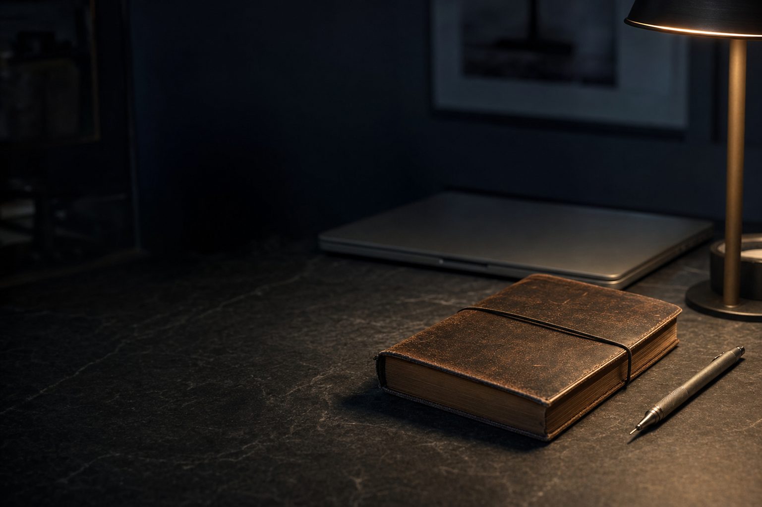 A leather notebook and a pen on a dark desk beside a closed laptop and a lamp.