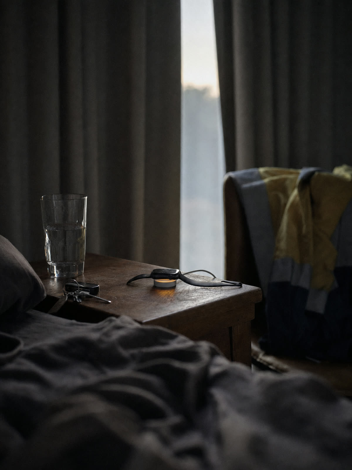 A darkened bedroom at dawn, with a glass of water and earplugs on the bedside table and the curtains drawn.