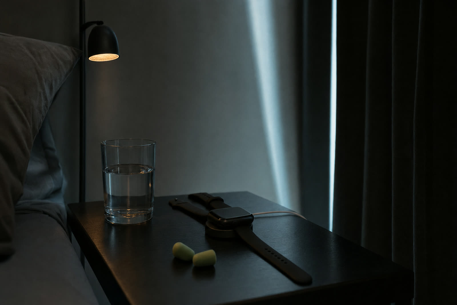 A smartwatch on its charger on a bedside table before dawn, beside a glass of water and earplugs.